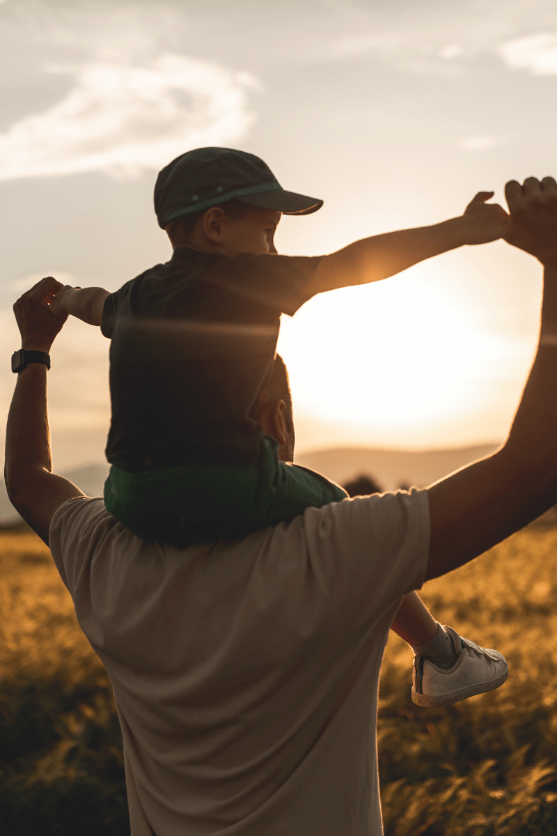 Das Bild zeigt einen Erwachsenen, der ein Kind auf seinen Schultern trägt. Sie genießen einen Sonnenuntergang auf einem Feld. Die Szene vermittelt Wärme, Geborgenheit und Freude in einem natürlichen Umfeld.